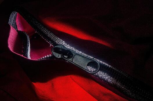 Close up on a zipper with a black slider on a red fabric garment, partly unzipped, revealing a pink lining. The zipper teeth are visible and the fabric is textured and shiny photo