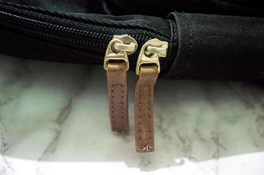 Close up on the zipper of a black bag featuring two brown leather zipper pulls. The bag is made of durable fabric, with a sturdy zipper mechanism. The background is marble photo