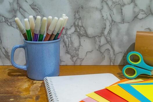 Close up shot displays a blue mug filled with colorful markers, a blank notebook, scissors, tape, and assorted color papers on a wooden table against a marble backdrop. Diy concept photo