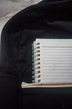 Close up on a spiral notebook with lined pages, partially inside a black backpack. The notebook is white and the backpack interior is beige, creating a monochromatic effect photo