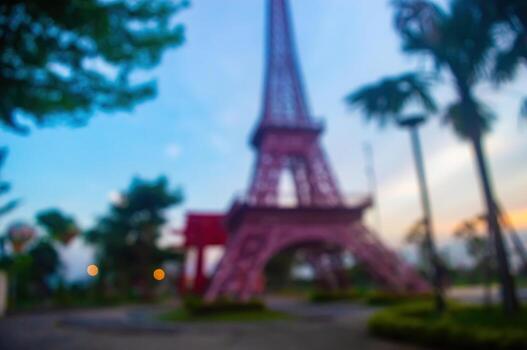Blurred outdoor scene with a miniature eiffel tower replica on the right, palm trees, and lush greenery against a soft blue and orange sky at dusk, creating a dreamy atmosphere. photo