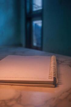 Close up of a spiral notebook with blank lined pages, resting on a table with a marble like surface, in a dimly lit room near a window, creating a sense of anticipation and creativity photo