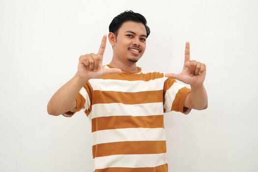 joven asiático hombre en blanco y marrón a rayas camiseta sonriente y enmarcado un rectángulo con ambos manos, en pie en contra un blanco fondo, como Si visualizante un foto o escena
