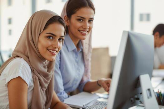 Two women in hijab are sitting at a desk with a computer monitor in front of them. They are smiling and seem to be working together photo