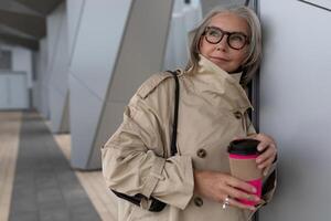 elegante mujer en neutral zanja Saco disfrutando café fuera de moderno edificio en un soleado día foto