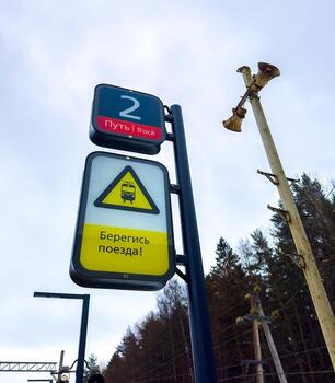 A low-angle shot of two signs on a platform. The top sign indicates Track 2 in Russian and English. Below it, a bright yellow triangular warning sign with a train icon warns Beware of the train in photo