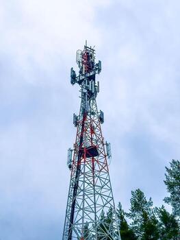 A dramatic, low-angle shot of a tall telecommunication tower with red and white sections, extending high into a cloudy sky. The steel lattice structure is framed by the tops of pine trees on either photo