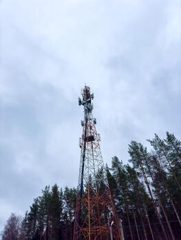 Dramatic, low-angle shot of a tall telecommunication tower with red and white sections, extending high into a cloudy sky. The steel lattice structure is framed by the tops of pine trees on either photo