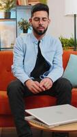 Indian man enters room starts typing on laptop browsing internet using apps sits on sofa. Happy young Hispanic guy working on computer in living room at home. E-learning browsing internet on notebook video