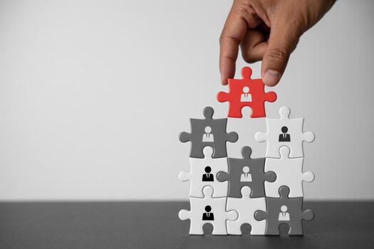 Hand placing red puzzle piece with a person icon on top of a stack of grey and white puzzle pieces representing a team, on a table, symbolizing human resource management photo