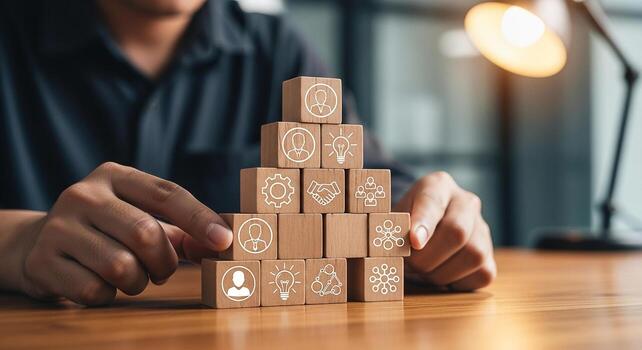 Person building a pyramid of wooden blocks with icons representing teamwork, strategy, and ideas in a modern office setting, symbolizing business growth and collaboration photo