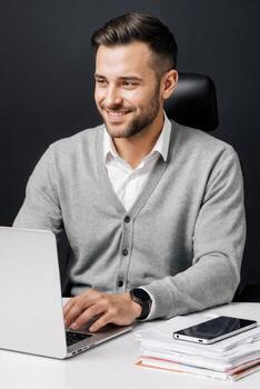 A smiling man in a sweater is sitting at a desk with a laptop photo