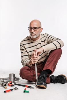 Experienced plumber demonstrates repair techniques while sitting amid tools and pipes in a workshop setting with a neutral background photo