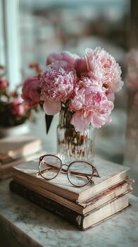 Beautiful Peonies in a Glass Vase on Stacked Books With Glasses on a Table by a Window photo