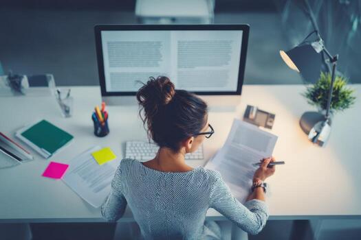 Businesswoman Working Late at Office Desk, Reviewing Documents and Typing on Computer photo