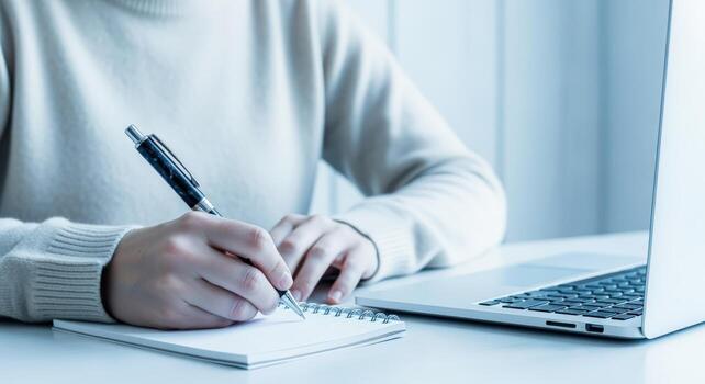 Close-up of Hands Writing in Notebook Beside Laptop Computer in Modern Office Setting photo