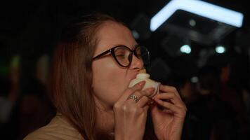 Young woman in glasses stands outdoors at night and enjoys eating mochi dessert with satisfaction, while blurry lights of concert or festival glow in background. video