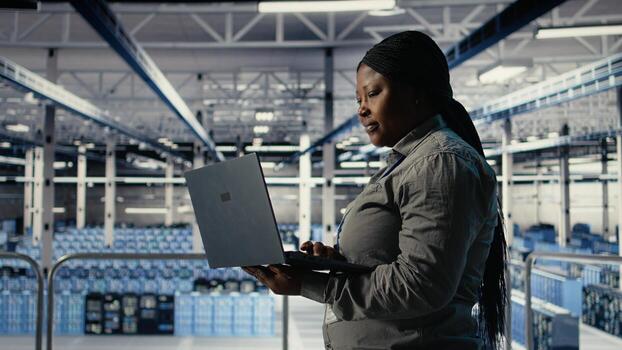 IT expert monitors server farm activity using laptop while walking in facility. African american woman in data center reviews rigs tests results to ensure systems are working correctly, camera A photo