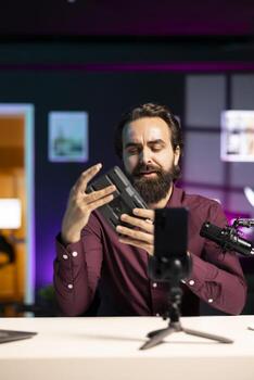 Tech specialist filming review of portable V mount battery using smartphone on tripod, feeling unconvinced of performance. Man analyzing lithium li-ion charging gadget, skeptical of quality photo