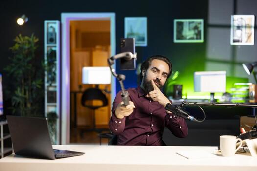 Online star talking in front on smartphone attached to selfie stick in green neon lit studio, discussing with viewers. Man using phone camera to record in apartment for social media photo