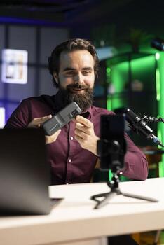 Joyful tech expert filming review of portable V mount battery using cellphone on tripod, happy with increased specs. Man excited about lithium li-ion charging gadget performance, endorsing it photo