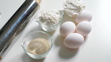 Flat lay of baking tools and ingredients for making bread on a white background, rolling pin, whisk, measuring cups, flour, yeast, and eggs video