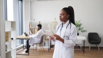 Female adult healthcare professional dressed in white coat using digital tablet in office setting. Office environment includes another healthcare worker seated in background working on computer. video