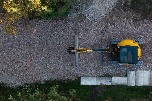 Yellow excavator on gravel surface with extended arm, bordered by grass and trees on the left and concrete slabs forming a pathway to the right. photo
