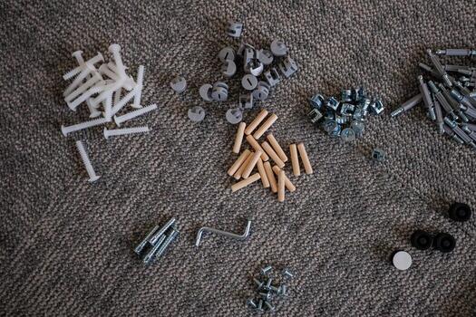 An assortment of hardware components, including screws, dowels, hex nuts, and an Allen wrench, neatly arranged on a textured carpet surface. photo