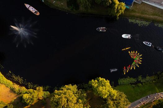 Aerial view of a waterway in Riga, Latvia, with colorful kayaks in a circular pattern, a fountain on the left, boats, trees, and a grassy bank visible. photo