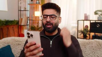 indisk man på Hem öppnas ögon bred och hissar ögonbryn medan ser på smartphone med chockade Lycklig reaktion. arab kille på soffa gester Wow med hand, överraskad förbi oväntat Bra meddelande. video