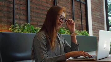 un mujer mira agotado mientras trabajando en frente de su computadora portátil, tomando apagado su lentes y masajear su frente. video