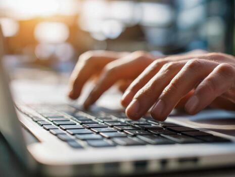 Hands of an adult person typing on laptop computer keyboard in a bright workspace with soft natural sunlight reflecting on the keys and background blur photo