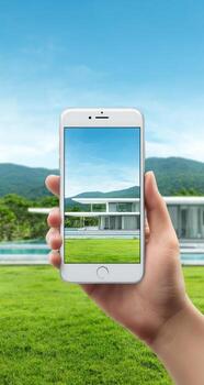 A hand holding a smart phone with a landscape view of a modern house in the background photo