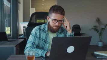 escritório trabalhador com Óculos e barba concentrando em computador portátil tela, pensativamente tocante dele cabeça enquanto trabalhando dentro moderno escritório ambiente. complexo trabalhos às computador dentro escritório video