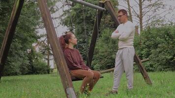 en par är utgifterna kvalitet tid tillsammans i en parkera. ett person sitter på en gunga medan de Övrig står närliggande, skapande en lugna och lekfull atmosfär omgiven förbi träd video