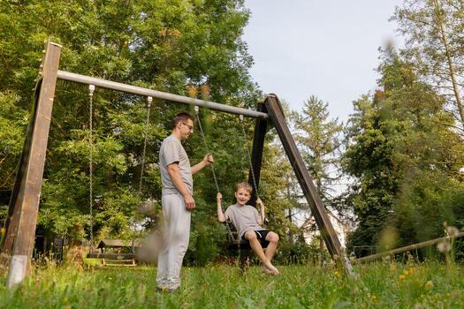 A father pushes his smiling child on a swing, both wearing matching gray outfits. They share joyful laughs in a beautiful park setting, filled with trees and greenery photo