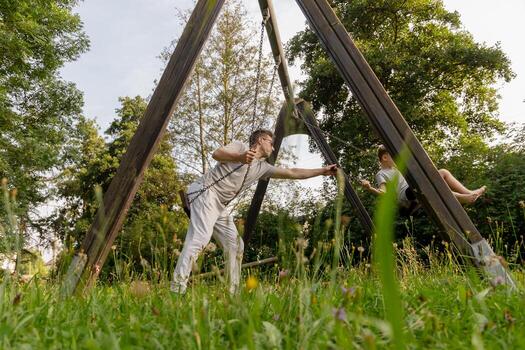 A joyful father pushes his child on a swing, both smiling and laughing. They are at a playground filled with green grass and trees, enjoying a warm afternoon together photo