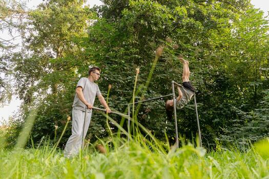 A father guides his child as they engage in playful activities at a playground. The child attempts to perform a handstand while surrounded by vibrant trees and grass photo