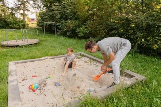 A father and his young child play together in a sandbox, creating shapes and using colorful toys. They are enjoying a sunny day in a park filled with trees and vibrant nature photo