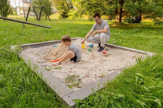 A father and his young child enjoy quality time together at a sandbox in a serene playground setting. Lush greenery surrounds them as they play and create memories photo