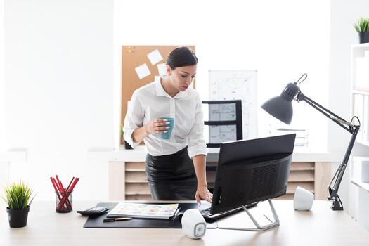 A young girl standing in the office, holding a Cup and typing on the keyboard. photo