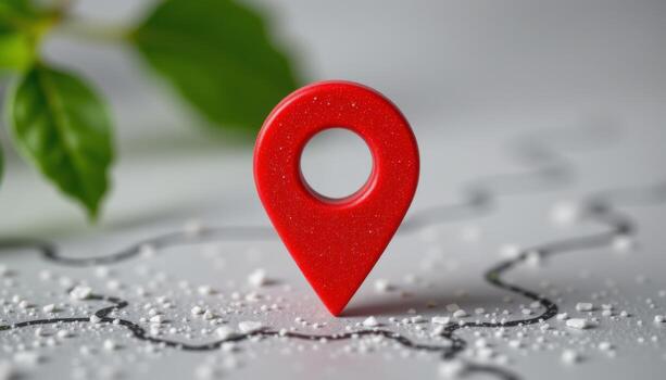 Vibrant red map pin icon standing on a light textured surface with a winding black path Blurry green leaves appear in photo