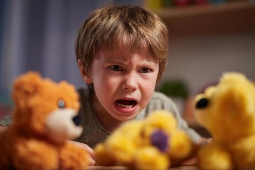 A child shows strong emotions while interacting with plush toys on a wooden table. The warm indoor lighting adds to the homey atmosphere, highlighting his expressions photo