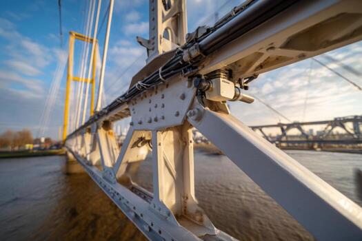 Detailed perspective of a suspension bridges metal framework and cables, highlighted by warm sunset light. The scene captures the serene river below and the soft clouds above photo