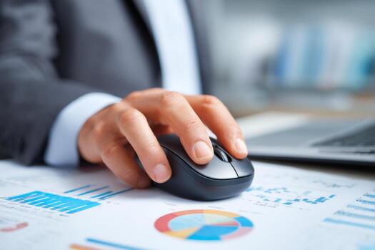 A business professional focuses on the computer mouse while reviewing financial reports spread out on a desk in an office setting. The workspace is well-organized with a laptop and charts photo