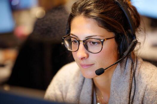 A woman with glasses works diligently at her desk in a call center. She wears a headset and focuses on her computer screen while providing customer service photo