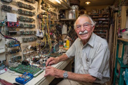 An elderly man focuses intently on assembling circuit boards in a cluttered workshop filled with an array of tools and electronic components. His dedication to his craft is evident photo