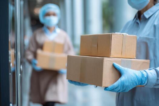 Two delivery workers are shown carrying cardboard boxes in an urban setting. They are wearing protective masks and gloves, focusing on their task during daytime photo