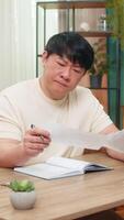 asiatisk man Sammanträde på Hem tabell rynkar pannan medan kontroll verktyg räkningar, skrivning anteckningar i anteckningsbok med fokus. kinesisk kille visas djup i trodde, beräknande försiktigt och analyserar finansiell detaljer. video